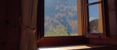 Una habitación con un ventanal amplio, cortinas y vistas a una cadena montañosa boscosa.