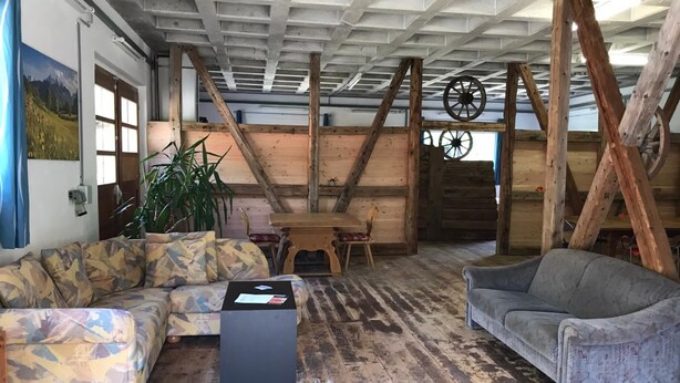 Ein rustikales Blockhaus-Interieur mit Holzbalken, einem Holztisch, zwei Sofas und einem großen Fenster.