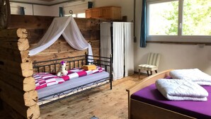 Ein rustikaler Raum mit einem Holzbettkasten, einem Einzelbett mit violettfarbener Matratze, einem Sessel und einem Fenster mit Vorhängen.