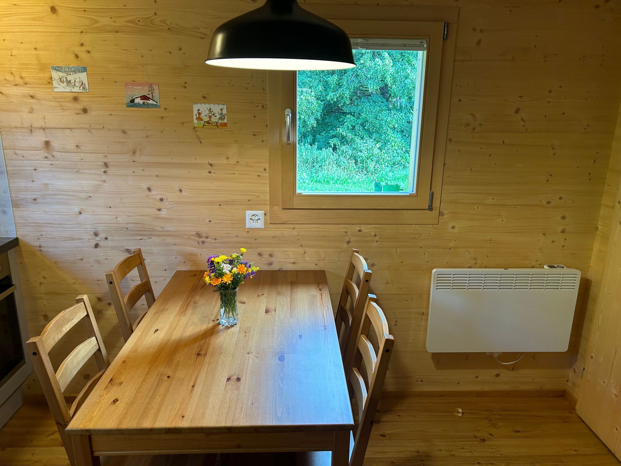 A wooden dining room with a table, chairs, a vase with flowers, a window with a view of greenery, and a wall-mounted heater.