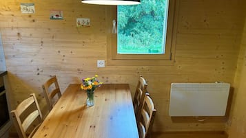 A wooden dining room with a table, chairs, a vase with flowers, a window with a view of greenery, and a wall-mounted heater.