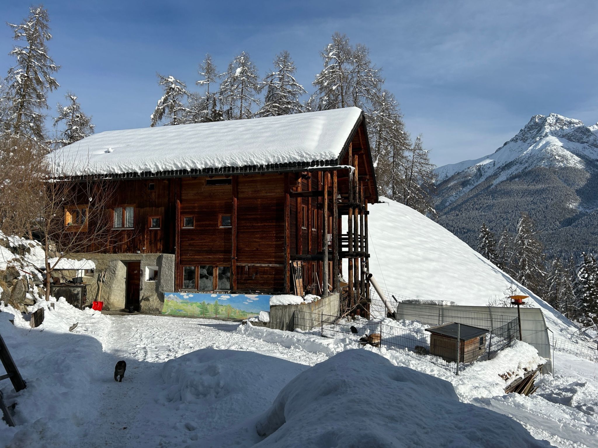 Una cabaña de madera con techo cubierto de nieve, rodeada de árboles y montañas nevadas.