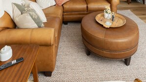 Una sala de estar con un sofá de cuero marrón, un reposapiés redondo, una mesa de centro de madera, una lámpara y vistas al exterior a través de grandes ventanales.