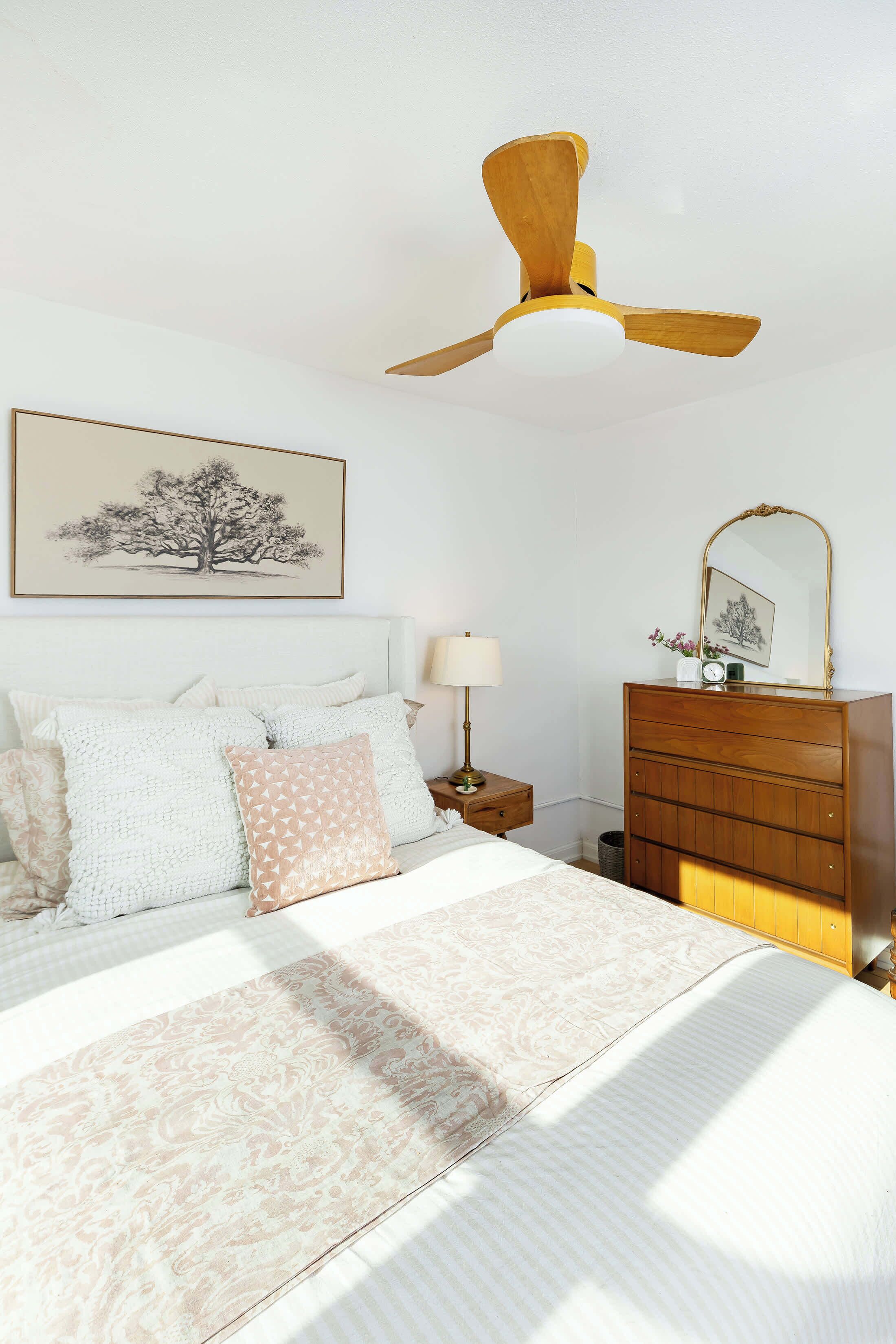 Een slaapkamer met een bed, een houten dressoir, een nachtkastje met lamp en een plafondventilator, plus een ingelijst beeld aan de muur.