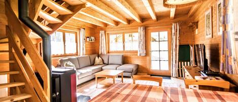 Interior de una cabaña de madera con escalera, sofá, mesa de comedor y televisor.