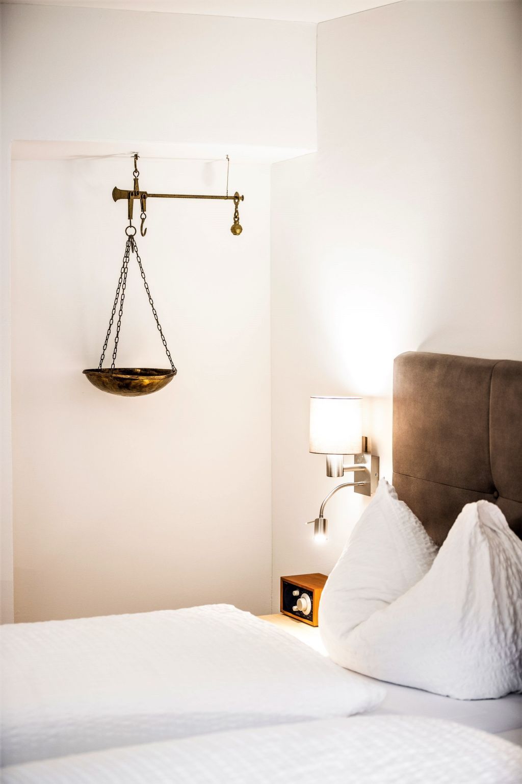 Quarto de hotel com cama, mesa de cabeceira com luminária e balança na parede.