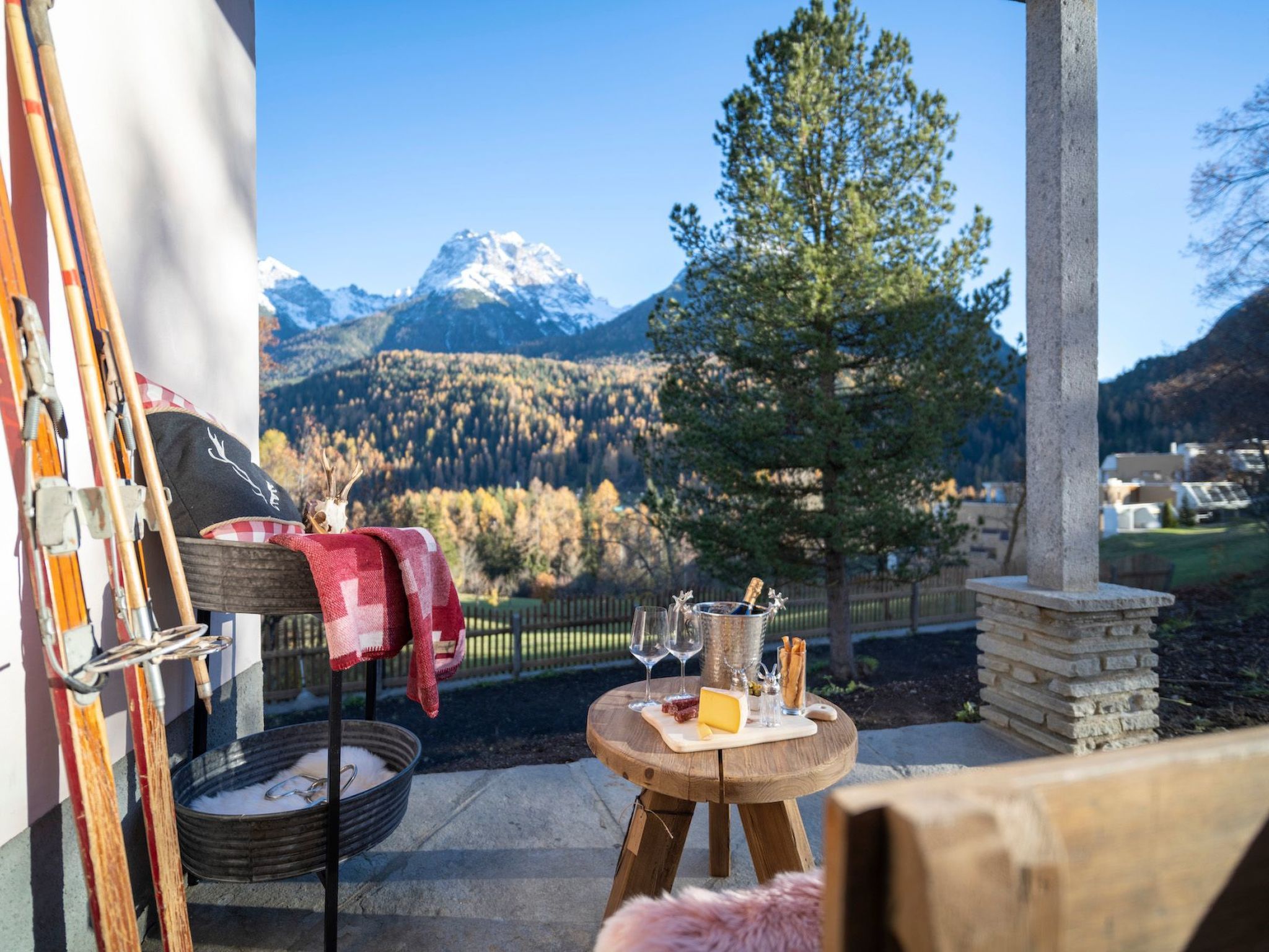A wooden table with a bottle of champagne, glasses, and a cheese board, set against a mountain backdrop.