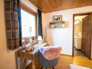 Une pièce chaleureuse avec un bureau en bois, une chaise recouverte d’un plaid en fourrure, une fenêtre avec des rideaux et une étagère ornée d’objets décoratifs.