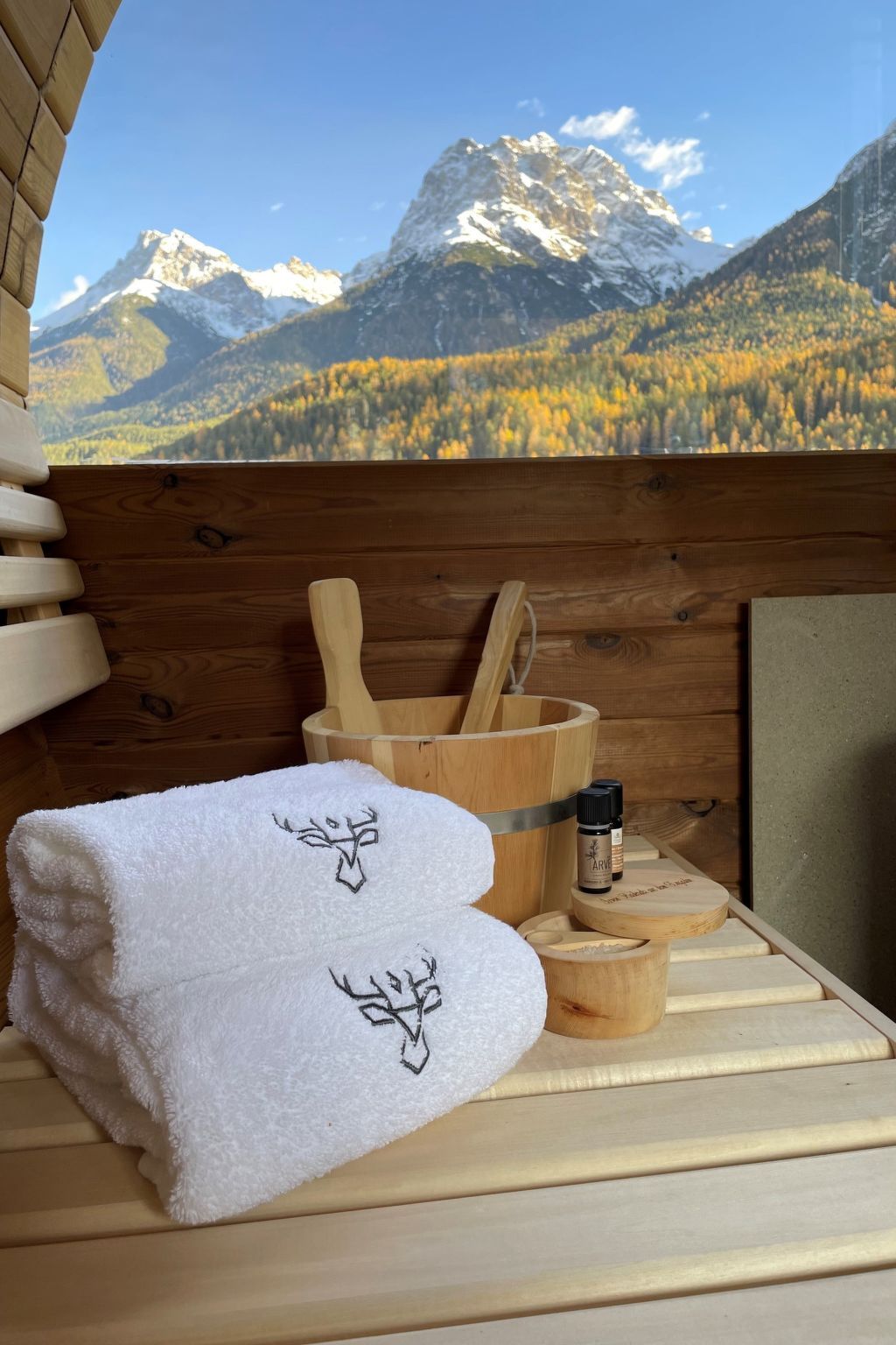 Interior de una cabaña de madera con vista a la montaña, una toalla blanca con diseño de árbol, un balde de madera y una botella de aceite esencial.