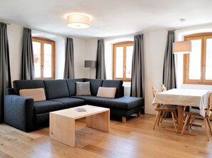 Ein modernes Wohnzimmer mit einem Ecksofa, einem Holztisch, einem Esstisch mit Stühlen und großen Fenstern mit Vorhängen.