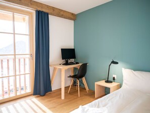 A bedroom with a bed, a desk with a computer, a chair, a nightstand with a lamp, and a view of the outdoors through a window with a curtain. - Chasa Muntanella, (Ftan) by Interhome (Ftan)