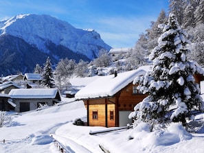 Un village de montagne enneigé, avec des maisons en bois, une montagne imposante en arrière-plan et un arbre recouvert de neige.