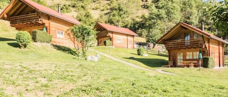 A row of wooden cabins with a grassy foreground and a forested hillside in the background.