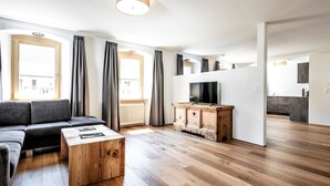Un salon moderne avec un canapé, une table basse, un téléviseur posé sur un meuble en bois, et une vue sur la cuisine.