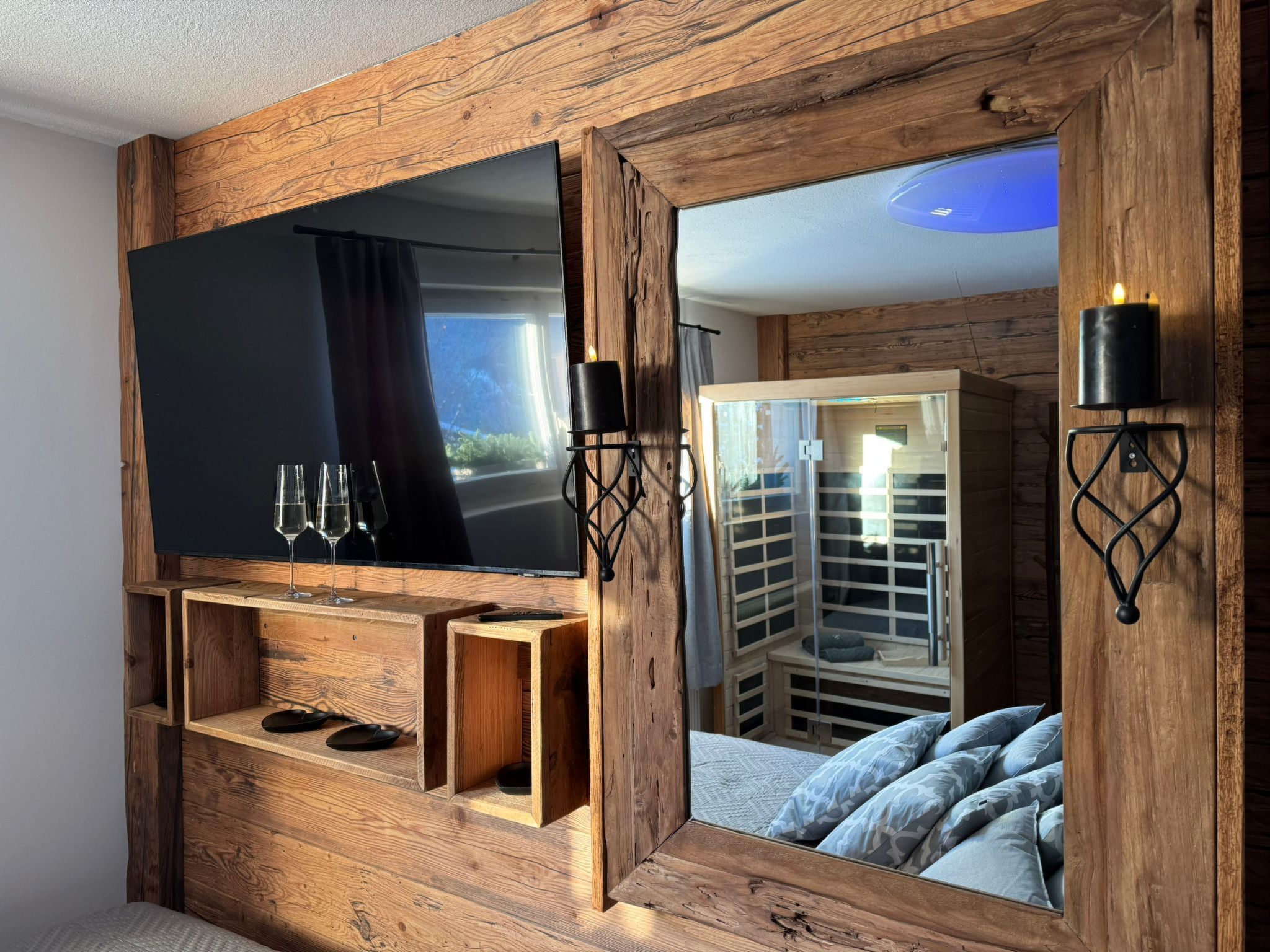 A wooden-framed TV with a shelf below, two wall sconces, and a mirrored wall reflecting a bed and a wardrobe.