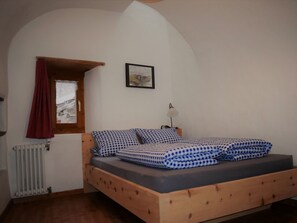 A bedroom with a wooden bed, checkered bedding, a window with a red curtain, a radiator, and a framed picture on the wall. - Agrotourismus Pütschai Josom, (Sta. Maria - Santa Maria Val Müstair) by Interhome (Sta. Maria GR)