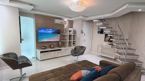 Un soggiorno moderno con una grande televisione a schermo piatto, un divano marrone, una scala e un lampadario.