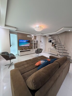 Ein modernes Wohnzimmer mit großem Flachbildfernseher, braunem Sofa, einer Treppe und einer Kristallleuchtre.