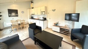 A modern living room with a black coffee table, two sofas, a dining area with a wooden table and chairs, a kitchen with white cabinets, and a mounted TV. - Bungalowpark Simpelveld - Schakel 6 by Interhome (Simpelveld)