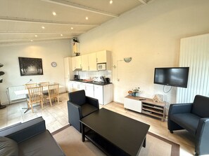 Un salon moderne avec une table basse noire, deux canapés, un coin repas avec une table et des chaises en bois, une cuisine avec des meubles blancs et un téléviseur fixé au mur.
