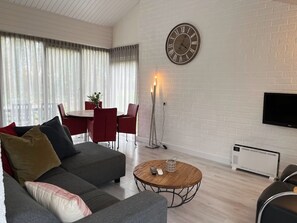 Ein modernes Wohnzimmer mit einer Couch, einem runden Couchtisch, einem Essbereich mit Stühlen, einer Wanduhr und einem Flachbildfernseher.