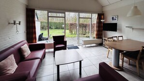 Ein Zimmer mit schrägem Dach, einer Couch, einem Tisch und Blick auf einen Garten durch große Fenster.