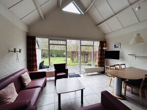 A room with a sloped ceiling, a sofa, a table, and a view of a garden through large windows. - Bungalowpark Simpelveld - Delta 154 by Interhome (Simpelveld)
