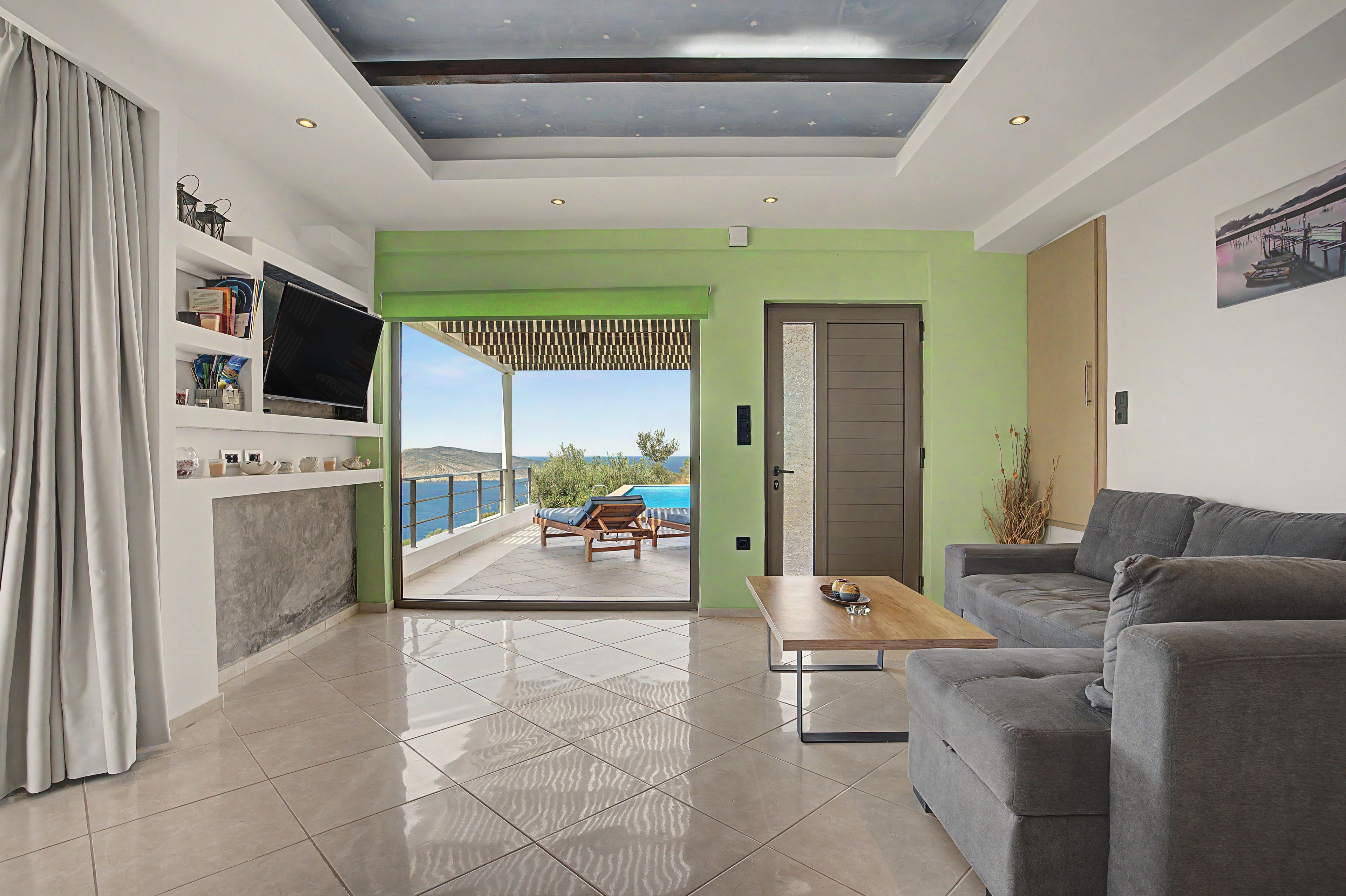 Una sala de estar moderna con un sofá, una mesa de centro y un televisor sobre una estantería. Hay una puerta que da a un balcón con vista al mar.