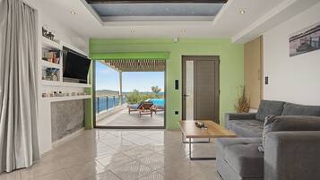 Una sala de estar moderna con un sofá, una mesa de centro y un televisor sobre una estantería. Hay una puerta que da a un balcón con vista al mar.