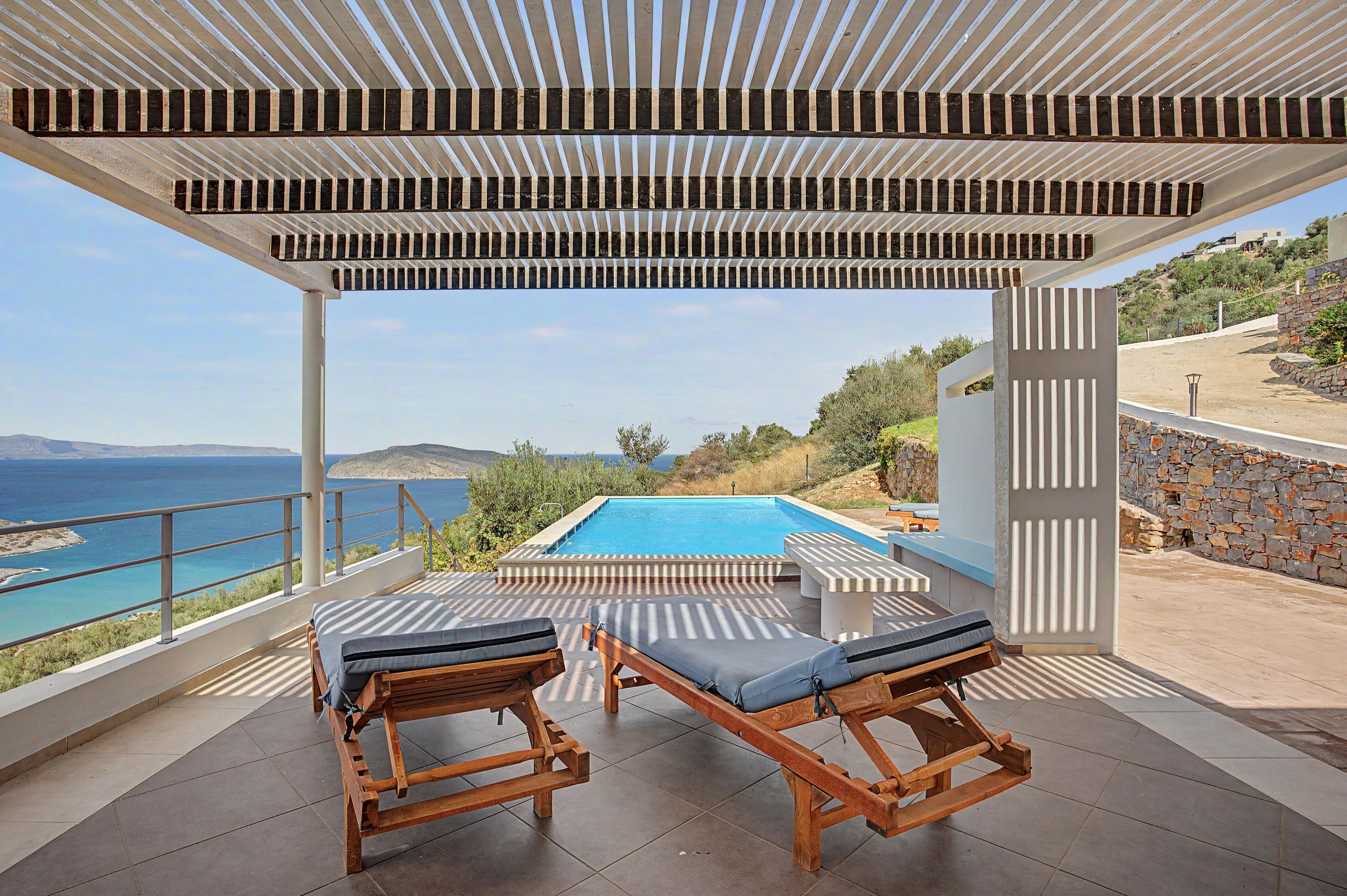 A covered outdoor area with a pool, two lounge chairs, and a view of the sea.