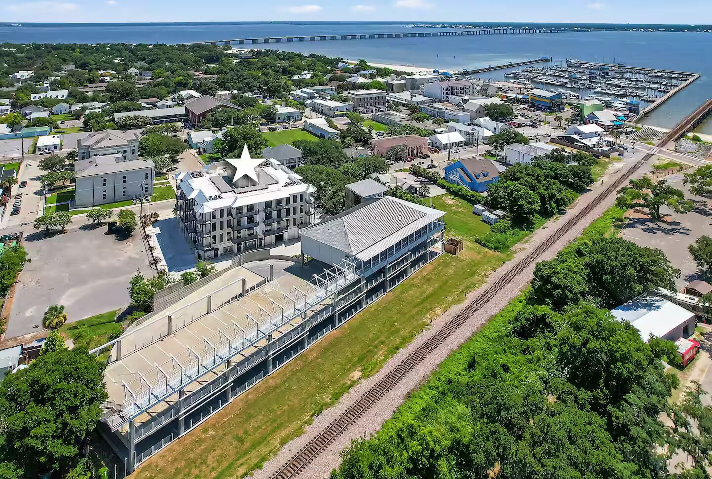 Vista aérea de un pueblo costero con un edificio destacado que tiene una gran estrella en el techo, un puerto deportivo y una vía ferroviaria que discurre paralela a la orilla.