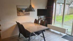 Uma sala de jantar com mesa, cadeiras, luminária pendente, televisão de tela plana e janela com cortinas.
