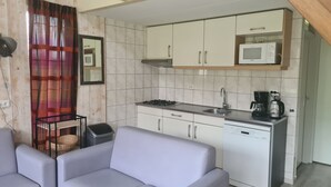 Cozinha compacta com armários brancos, micro-ondas, fogão e cafeteira. Na sala de estar, um sofá cinza e uma cesta de vime.