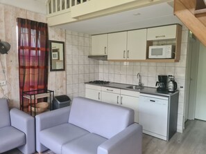 Eine kompakte Küche mit weißen Schränken, einer Mikrowelle, einem Herd und einer Kaffeemaschine. Im Wohnbereich befindet sich eine graue Couch und ein Korbwannenmöbel.
