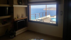 Ein Zimmer mit großem Fenster mit Blick auf einen Pool und das Meer, einer Uhr an der Wand und einem Fernseher auf einem Regal.