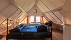 Ein Schlafzimmer im Loft-Stil mit schrägem Dach, einem Bett mit blauer Bettwäsche, einem Sessel und einem Fenster mit Vorhängen.