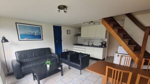 Un salón con un sofá, una mesa de centro y una mesa de comedor con sillas. Se observa una cocina al fondo con armarios y una estufa. Una escalera es visible a la derecha.