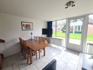 A dining area with a table and chairs, a television, and a view of a green lawn through a sliding glass door. - Bungalowpark Simpelveld - Delta 109 by Interhome (Simpelveld)