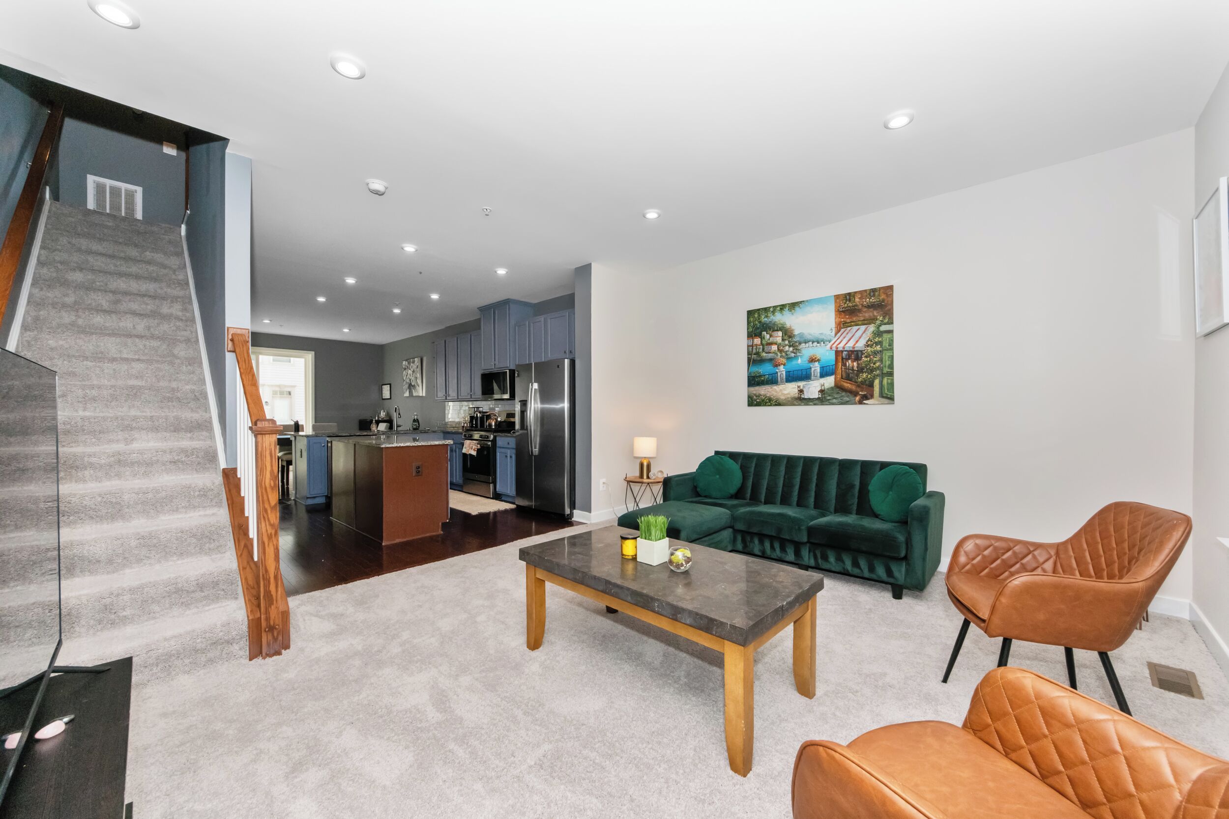 Un salon moderne avec un canapé, une table basse et des fauteuils, un escalier et une cuisine en arrière-plan.