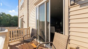 Una terraza de madera con mobiliario de mimbre, una puerta corrediza de vidrio y una pequeña zona de cocina al aire libre.