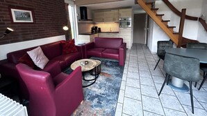 Ein Wohnzimmer mit rotem Sofa, Couchtisch und Esstisch mit Stühlen. Im Hintergrund befindet sich eine Küche mit Schränken und Geräten. Eine Holztreppe führt auf eine obere Ebene.