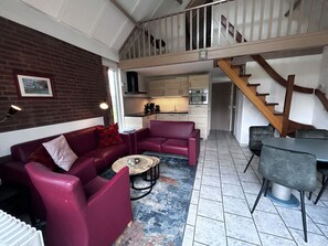 A living room with a red sofa, a coffee table, and a dining table with chairs. There is a kitchen in the background with cabinets and appliances. A wooden staircase leads to an upper level. - Bungalowpark Simpelveld - Delta 112 by Interhome (Simpelveld)