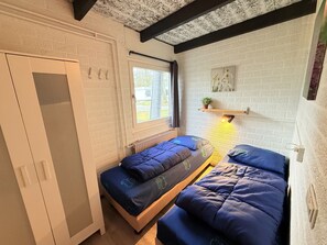 Ein Zimmer mit zwei Betten, einem Fenster, einer Heizung und einem Regal mit einer Pflanze.