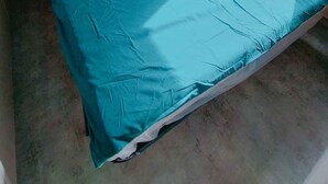 Een enkel bed met een blauwe matras, een houten hoofdbord, een plank boven het bed en een raam met gordijn.