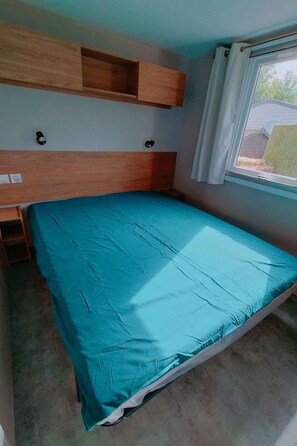 Ein Einzelbett mit blauer Matratze, einem hölzernen Kopfteil, einem Regal darüber und einem Fenster mit Vorhang.