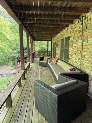 A covered porch with a stone wall, wooden deck, wicker furniture, and a bench. - Cozy Waterfront Log Cabin Retreat on a Quiet Creek (Mineral)