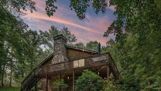 A cabin with a stone chimney, wooden deck, and a staircase leading to a balcony.