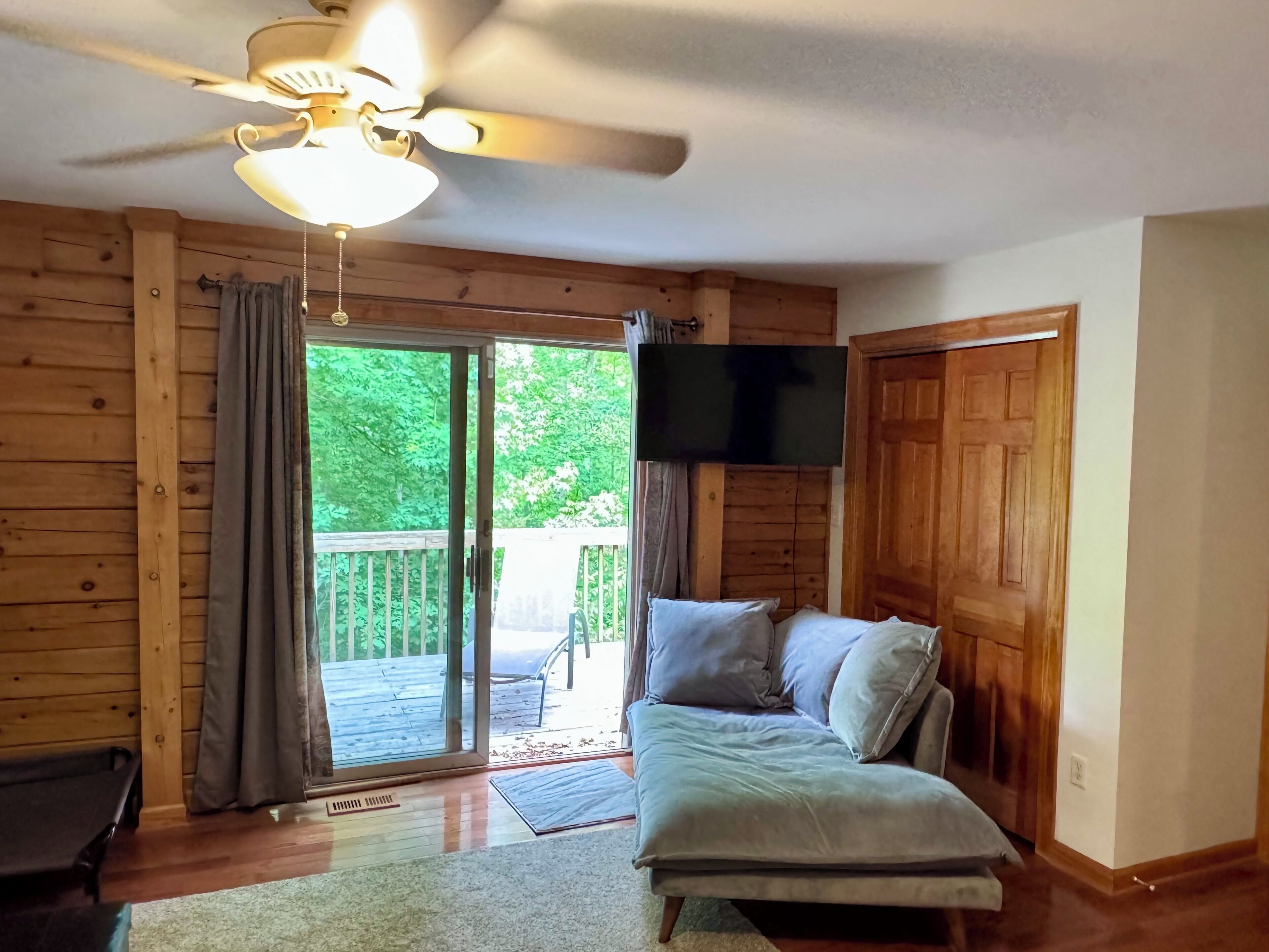 A room with a ceiling fan, a sliding glass door leading to an outdoor deck, a sofa with cushions, and a flat-screen TV mounted on the wall.