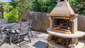 An outdoor grill with a stone structure, a table with chairs, and a wooden fence in the background. - Villa “My Little Castle Platja D'Aro” with Sea View, Balcony and Wi-Fi (Castell d'Aro)