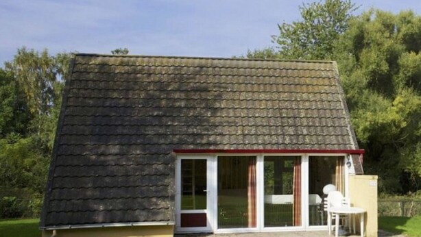 A single-story house with a sloped roof, a small porch with a table and chairs, and a pathway leading to the entrance. - Bungalowpark Simpelveld - Alpha 89 by Interhome (Simpelveld)